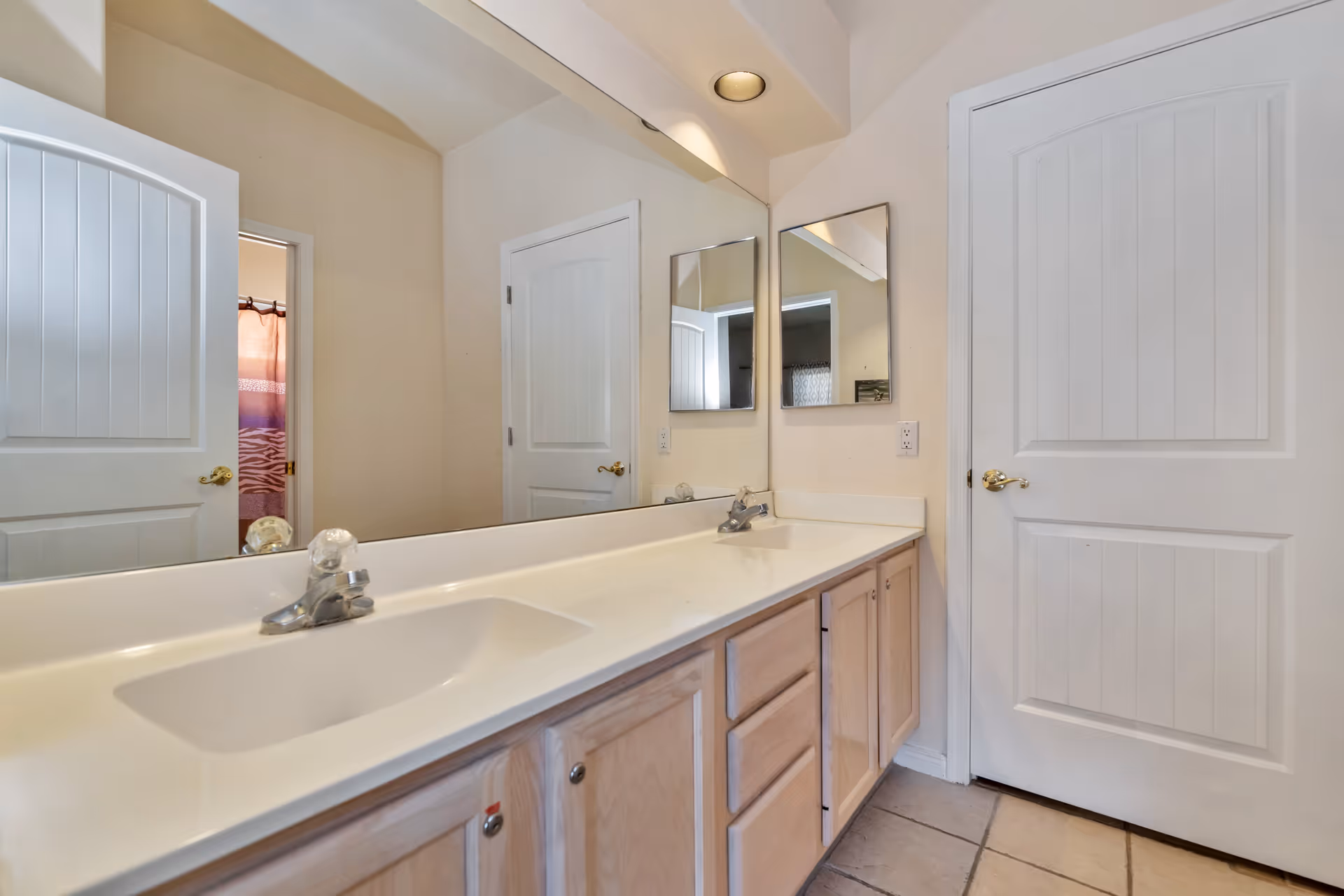 A bathroom interior featuring a long white countertop with two sinks and wooden cabinets underneath. There are two mirrors on the wall above the countertop, and two white paneled doors, one of which is slightly open revealing a shower curtain with a purple and pink pattern.