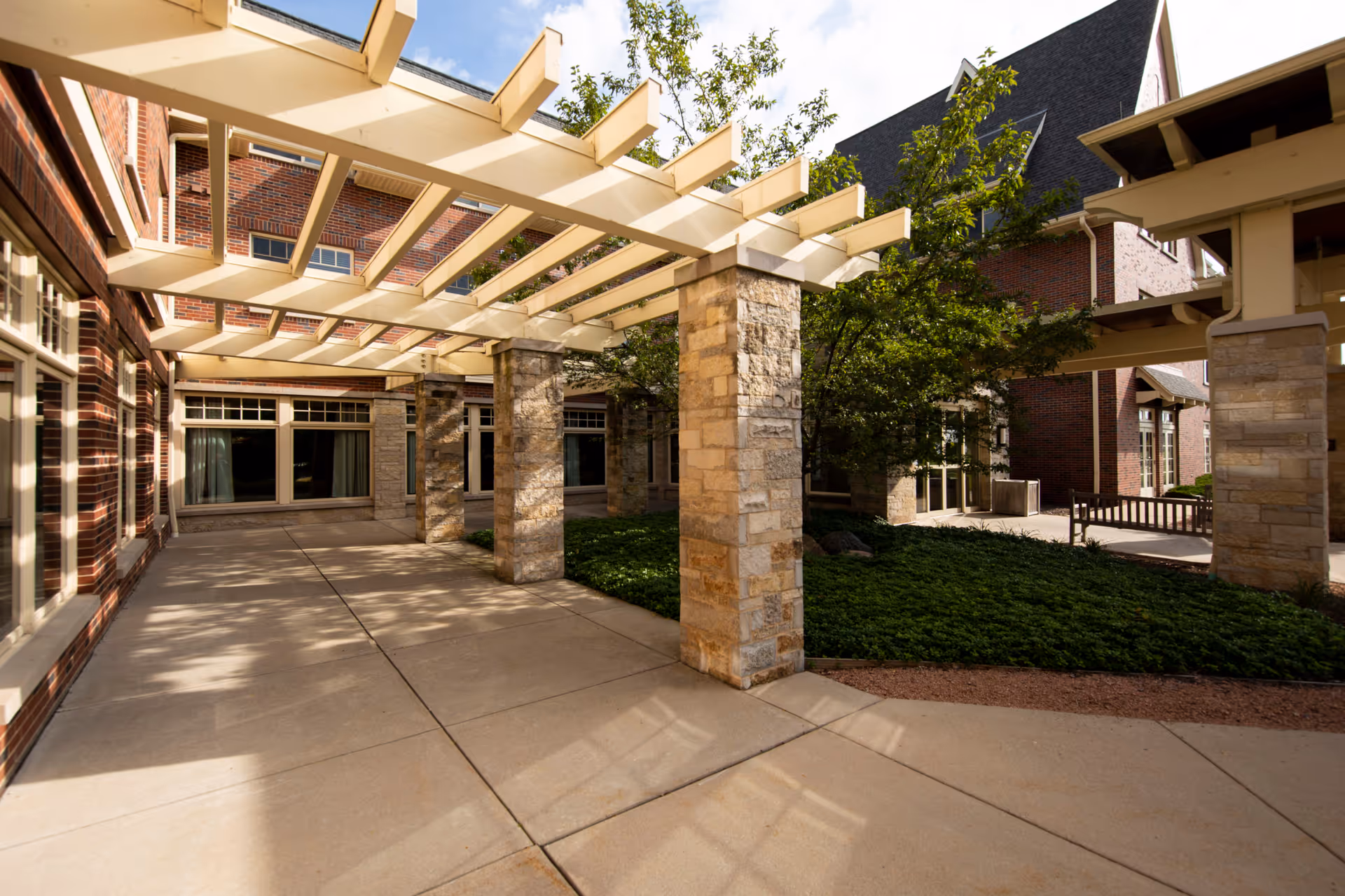 Sunlit courtyard with a pergola supported by stone pillars between brick buildings and landscaped greenery.