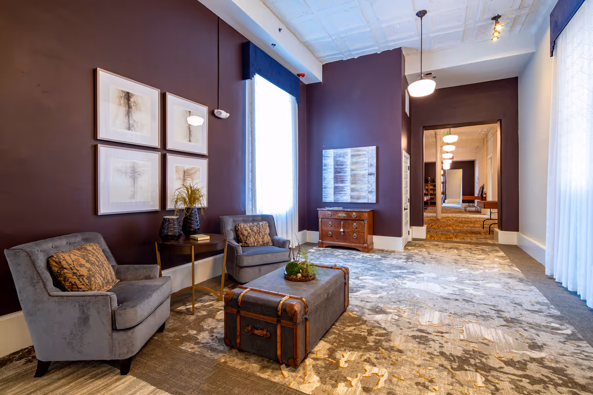 A stylish common seating area with two upholstered chairs, a trunk-style ottoman, artwork on purple walls, and a view into a hallway.