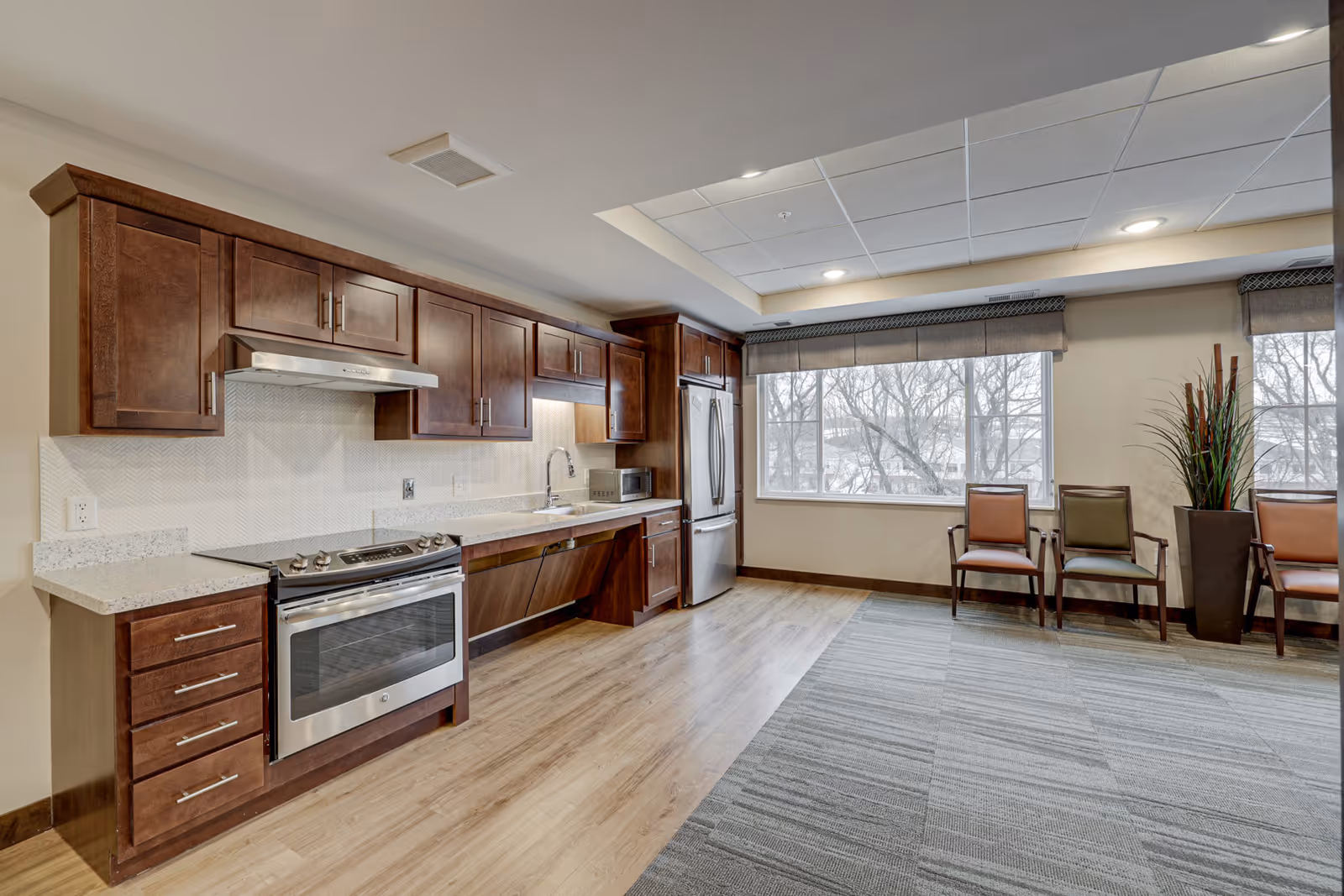 Bright communal kitchen and seating area with dark wood cabinets, stainless-steel appliances, and chairs by a large window.