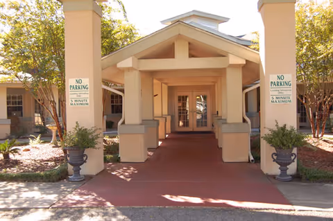 Covered entrance to a building with beige pillars and a red concrete walkway. Two signs on the pillars read 'NO PARKING Loading/Unloading Only 5 MINUTE MAXIMUM'. There are potted plants on either side of the entrance and trees in the background.