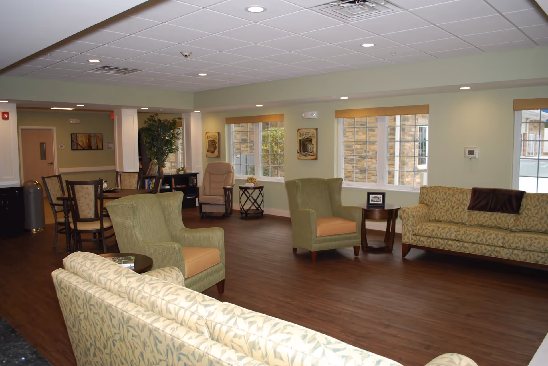 A spacious and well-lit living room area in a senior living facility with multiple upholstered chairs and sofas arranged around small tables. The room has large windows with wooden blinds, light green walls, and dark wood flooring. There is a dining table with chairs in the background and some framed artwork on the walls.