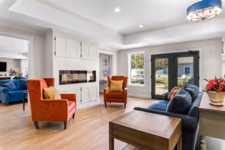A bright and spacious living room with light wood flooring, white walls, and recessed lighting. The room features a dark blue sofa, two orange armchairs with yellow cushions, a wooden side table, and a built-in white cabinet with a fireplace. Large windows and glass doors allow natural light to fill the space, and a modern ceiling light fixture is visible.
