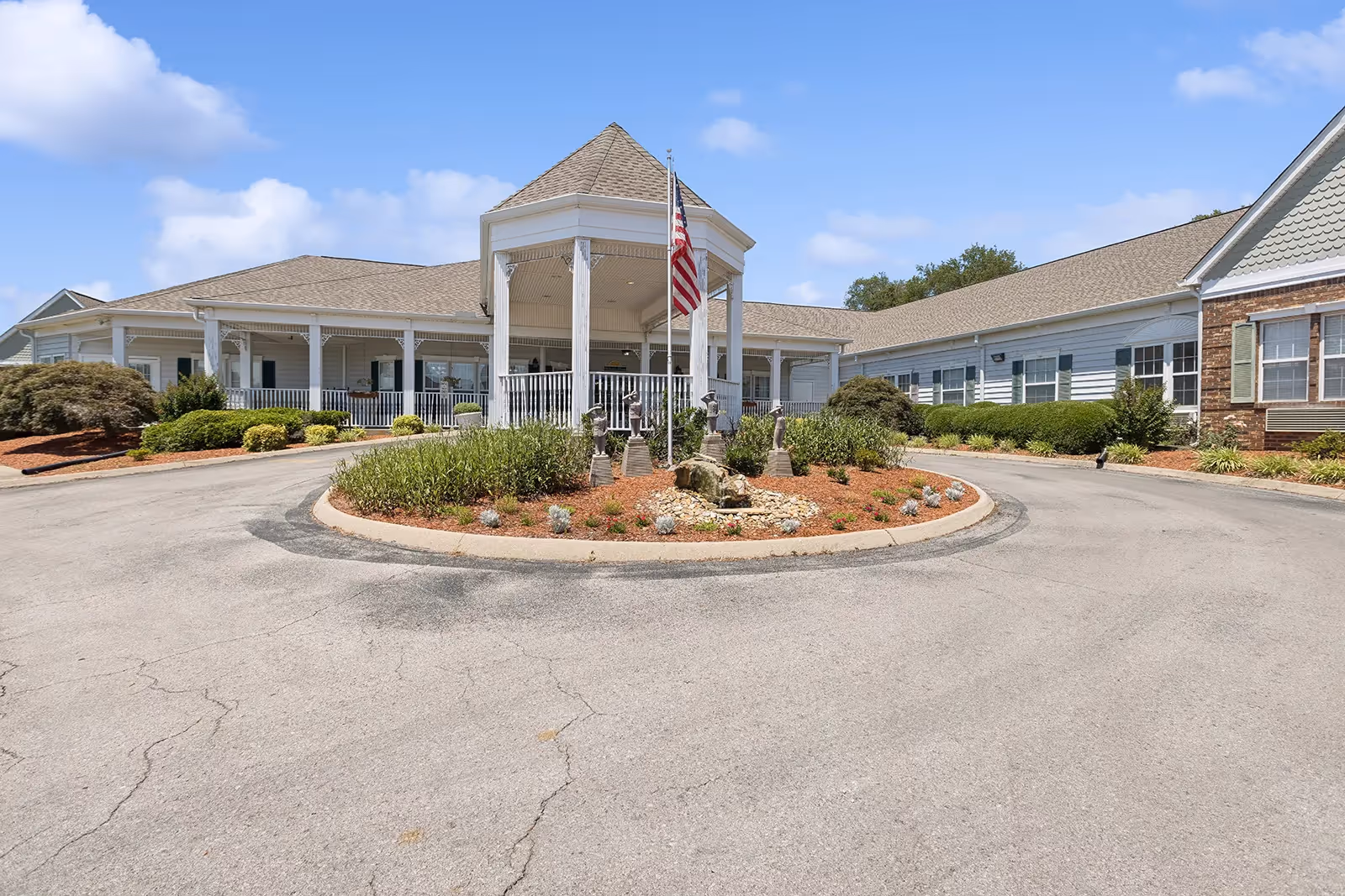 Front entrance of a senior living facility with a covered porte-cochère, American flag, circular driveway, and landscaped island.