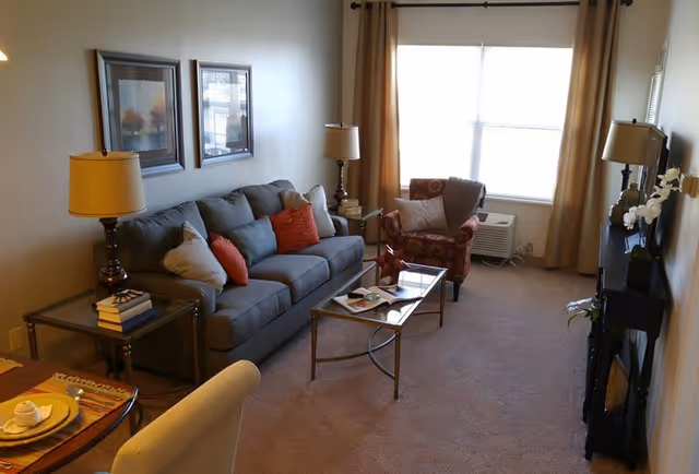 A cozy living room with a gray sofa adorned with orange and beige cushions, a glass coffee table with a book and decorative items, a patterned armchair near a window with beige curtains, two table lamps on side tables, framed artwork on the wall, and a TV on a stand with a small plant beside it.