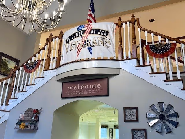 Interior view of a senior living facility with a staircase decorated for Veterans Day. A banner reading 'In Honor of Our Veterans' hangs on the railing, along with an American flag and patriotic bunting. Below the staircase is a sign that says 'welcome' with a quote. The walls are decorated with framed art and a metal windmill decoration.