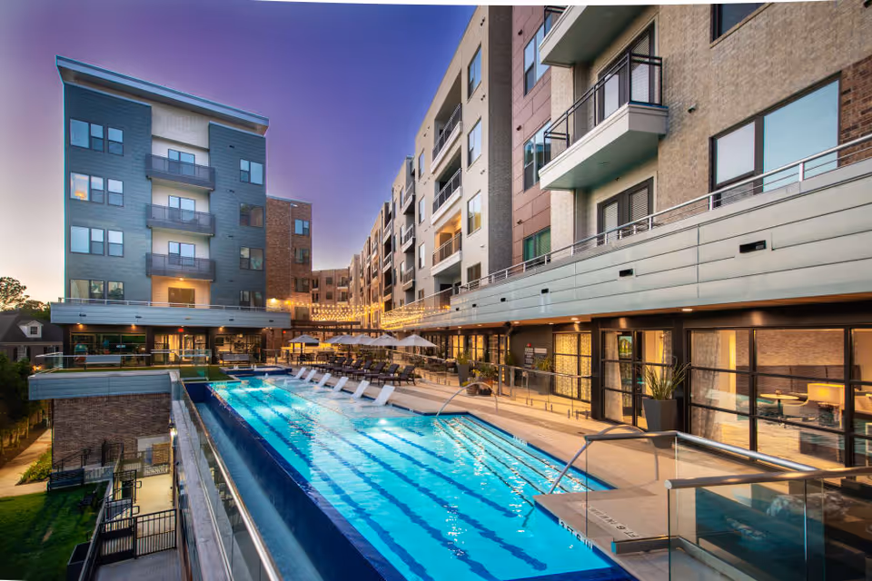 A modern multi-story residential building surrounding an illuminated outdoor swimming pool and lounge area at dusk.