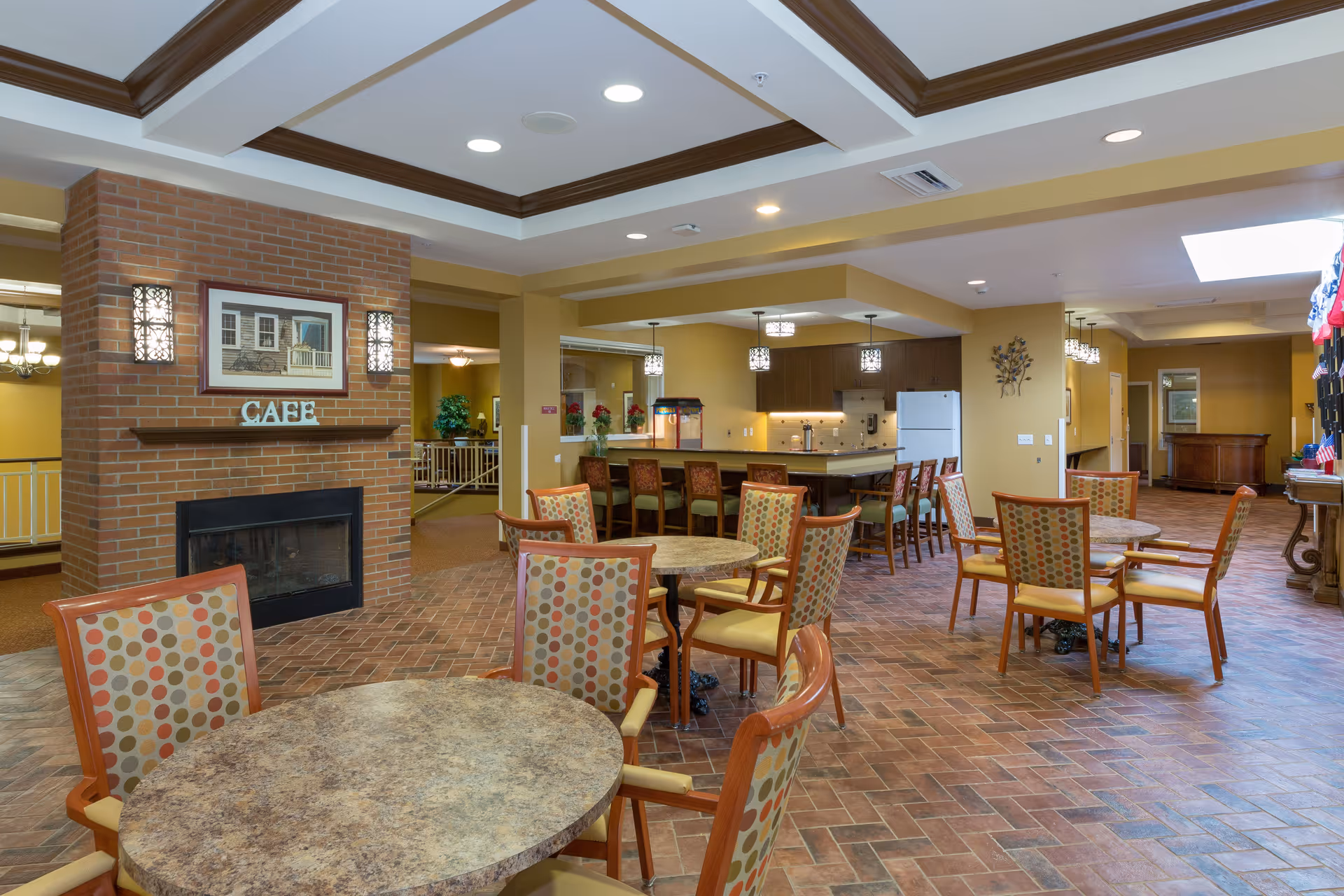 Interior view of a senior living facility cafe area with round tables and chairs featuring patterned upholstery. A brick fireplace with a framed picture and the word 'CAFE' sits on the left. In the background, there is a kitchen area with a refrigerator, cabinets, and a counter with bar stools. The room has warm lighting and a tiled floor.