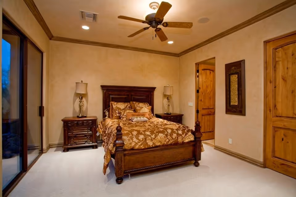A warmly lit bedroom featuring a wooden bed with an ornate headboard and footboard, dressed in a brown and gold floral patterned comforter and pillows. There are two matching wooden nightstands on either side of the bed, each with a decorative lamp. The room has beige walls, a ceiling fan with wooden blades, recessed lighting, and wooden doors. A large sliding glass door is visible on the left side of the room.