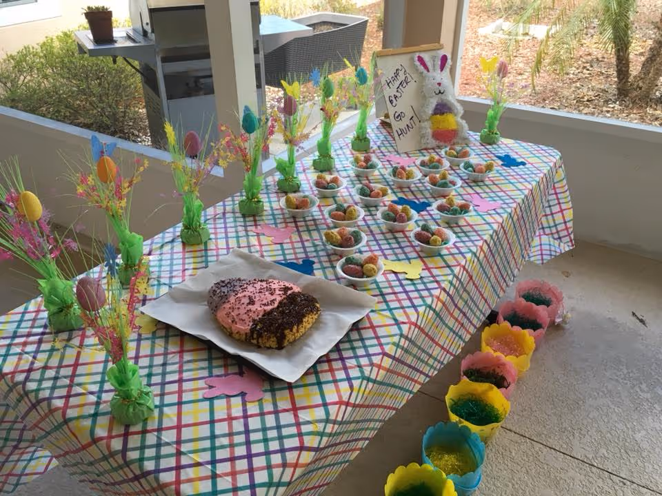 A table on a covered patio decorated for Easter with a plaid tablecloth holding small bowls of candy, spring floral picks, a large frosted pastry, and decorative baskets.