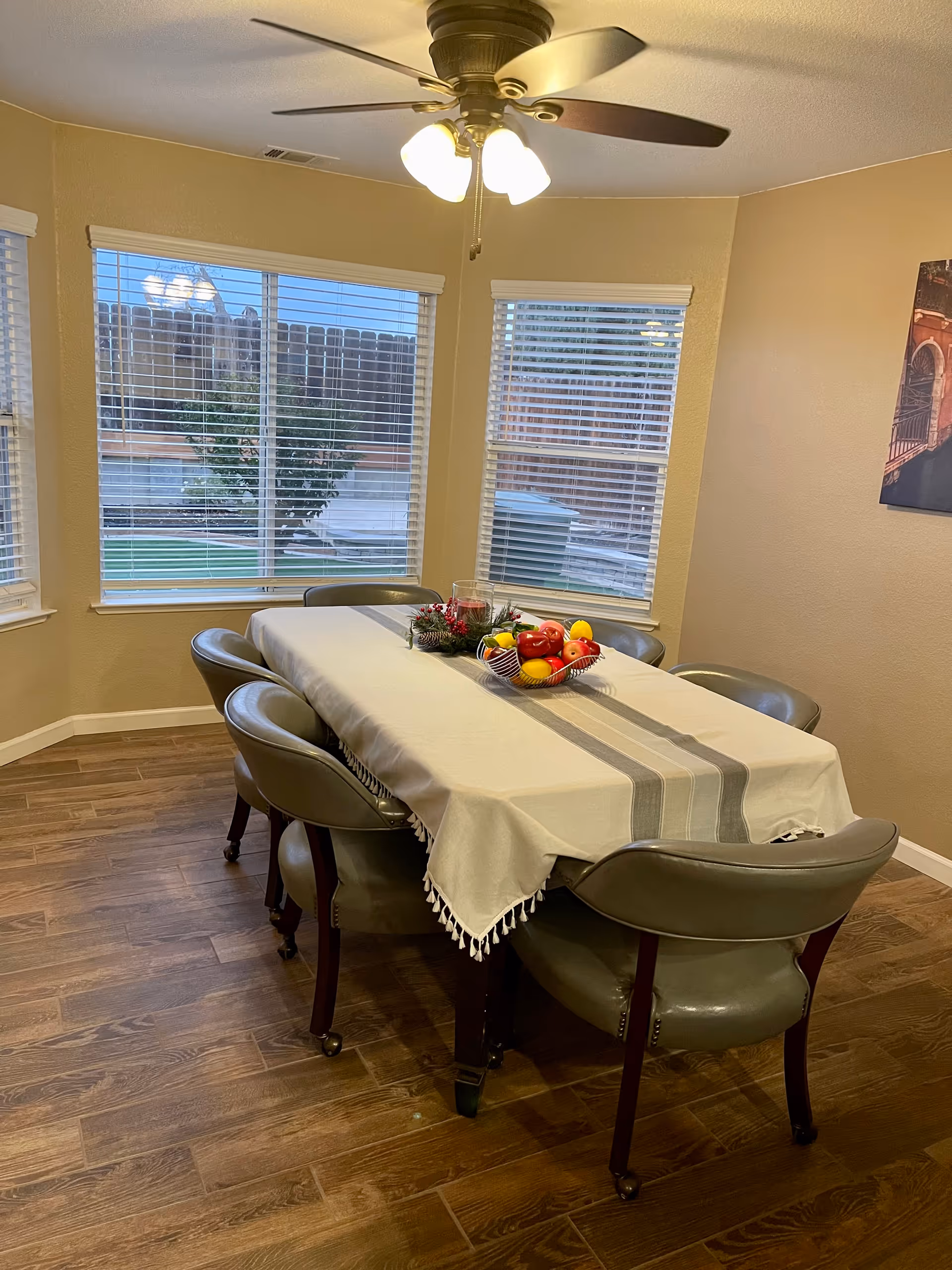 A dining room with a rectangular table covered with a white tablecloth featuring gray stripes. Six gray cushioned chairs surround the table. On the table, there is a decorative centerpiece with artificial fruit and greenery. The room has wooden flooring, beige walls, three large windows with white blinds, and a ceiling fan with lights.