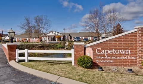 Exterior view of Capetown Senior Living facility showing a brick entrance sign with the facility name and description, a white fence, a driveway, and the building in the background under a partly cloudy sky.