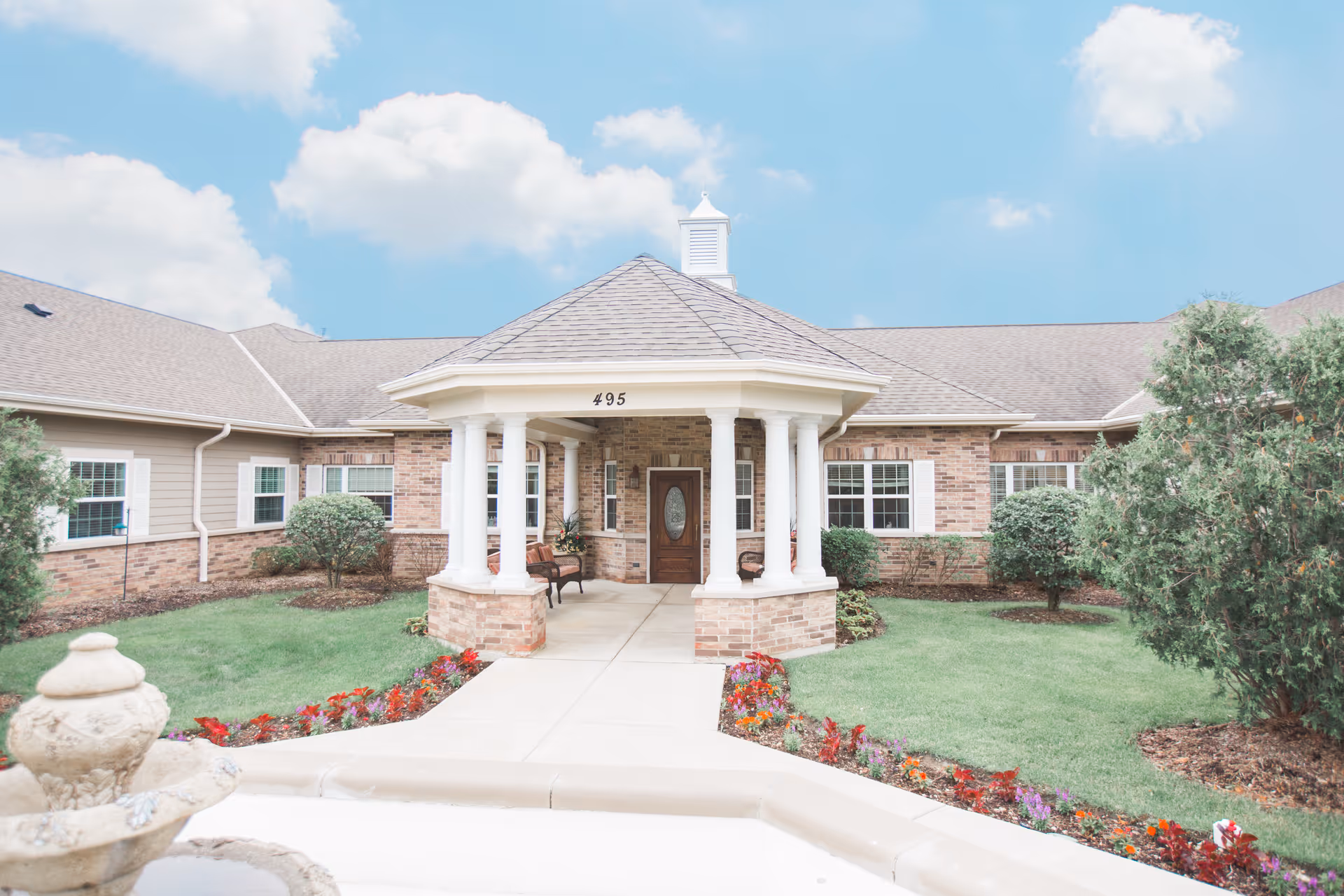 Front entrance of a single-story brick building with a covered porch supported by white columns, surrounded by green lawns, shrubs, and colorful flower beds under a partly cloudy blue sky.