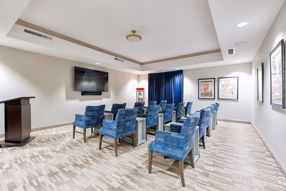 A small conference or presentation room with rows of blue patterned chairs facing a wall-mounted flat screen TV. There is a wooden podium on the left side and a popcorn machine in the back corner near dark blue curtains. The room has light-colored walls and carpet, with framed posters on the right wall.