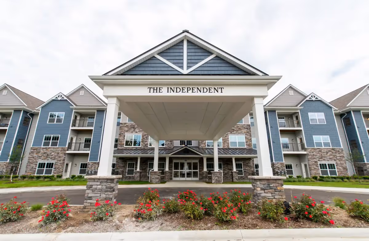 Front exterior view of a multi-story retirement living building named 'The Independent' with a covered entrance supported by white columns and landscaped flower beds with red flowers in front.