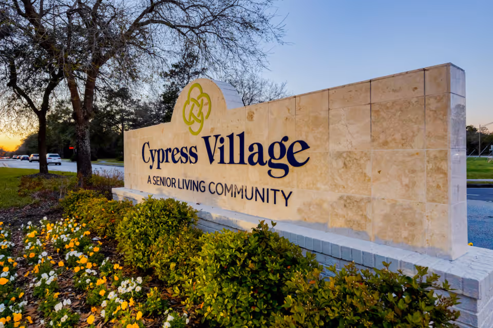 Stone sign for Cypress Village, a senior living community, surrounded by green bushes and yellow and white flowers, with trees and a road in the background at sunset.