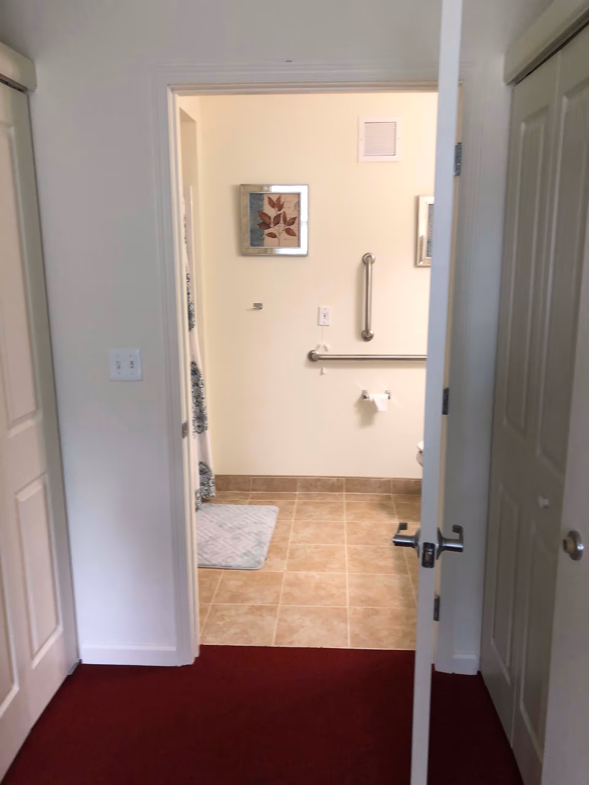 View through an open door into a bathroom with beige tiled floor, grab bars on the wall, a toilet paper holder, a framed picture, and a shower curtain partially visible on the left.