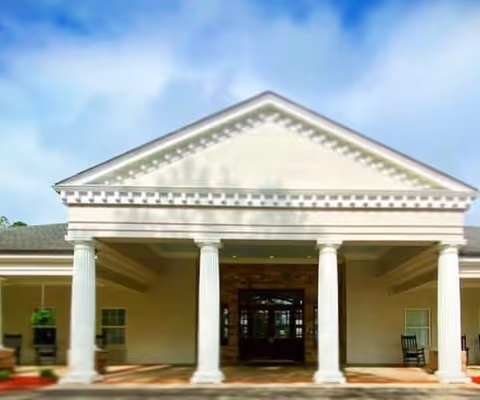 Front exterior view of a building with a classical architectural style featuring four white columns supporting a triangular pediment above the entrance. The building has a covered porch area with rocking chairs on either side and a clear blue sky in the background.