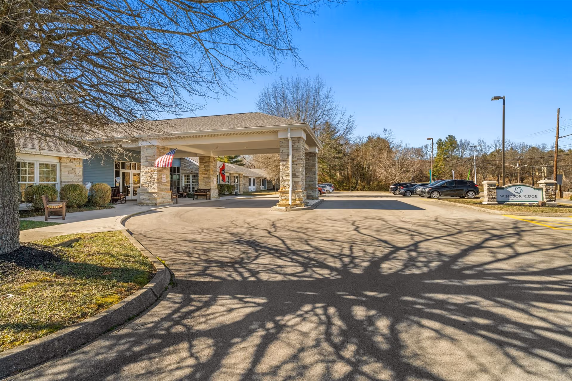 Exterior view of the Windsor Ridge senior living facility entrance with a covered drop-off area supported by stone pillars, an American flag and another flag hanging, parked cars in the parking lot, and leafless trees casting shadows on the pavement under a clear blue sky.