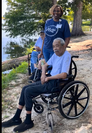 An elderly man in a wheelchair is outdoors near a body of water with trees in the background. A woman wearing a navy blue shirt with the word 'Nurse' on it stands behind him, smiling. Another elderly person in a wheelchair is visible further back along the path.