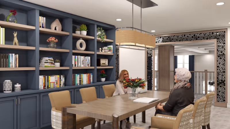 Two women sit at a long wooden table in a bright communal dining/library room with built-in bookshelves and a floral centerpiece.
