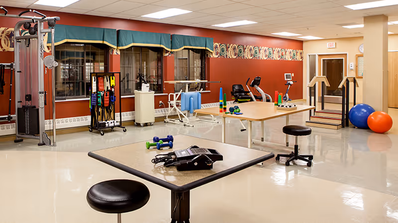 A spacious rehabilitation or physical therapy room with exercise equipment including a multi-station gym machine, stationary bike, therapy balls, dumbbells, resistance bands, and parallel bars. The room has a red accent wall with windows covered by green valances, beige tiled floor, and tables with stools.