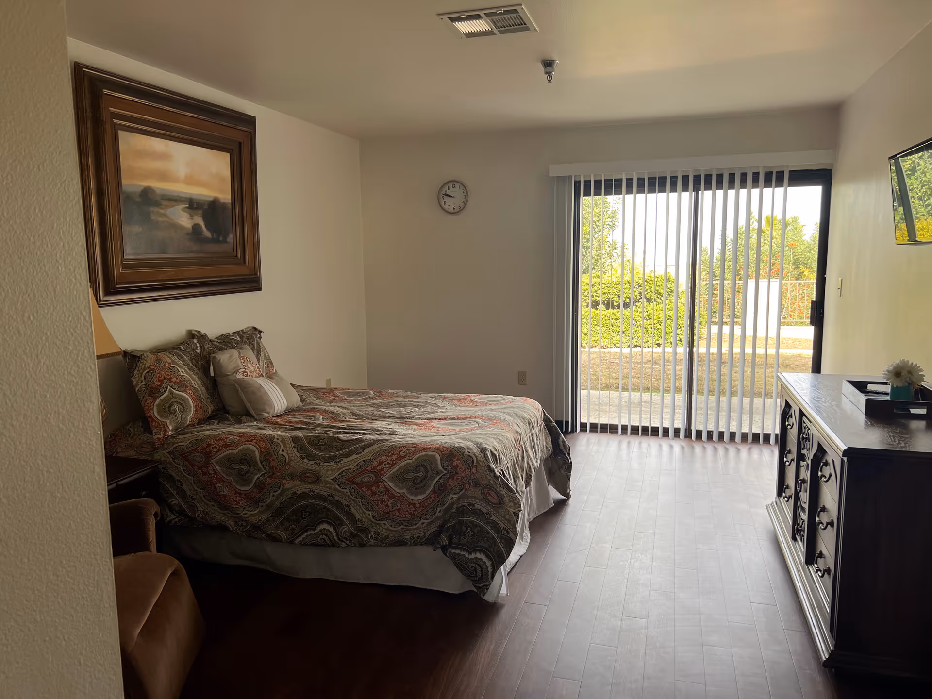 Sunlit bedroom with a bed, dresser, sliding glass doors with vertical blinds, and wall art.