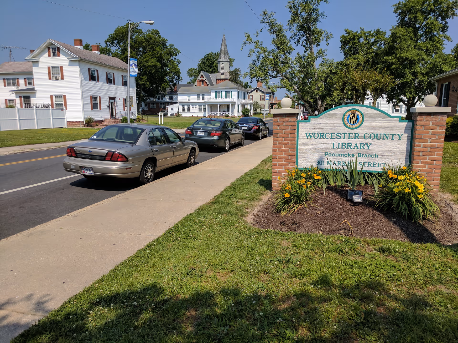 Street view with parked cars and a brick sign reading Worcester County Library Pocomoke Branch in front of houses and a church steeple.