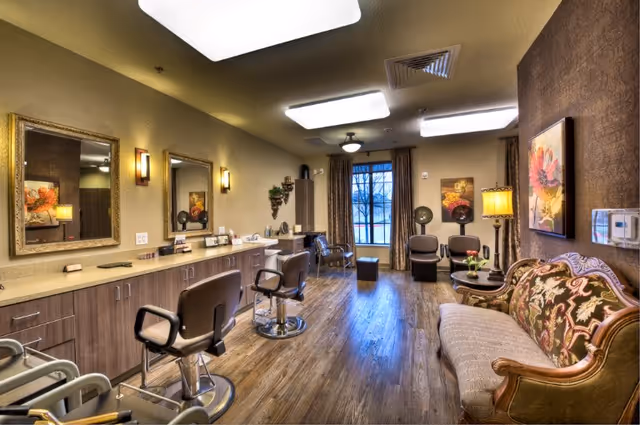 Interior view of a salon area in a senior living facility with three salon chairs in front of mirrors on the left side, wooden flooring, a vintage-style sofa on the right, and a window with curtains at the far end. The room is warmly lit with ceiling lights and wall sconces.