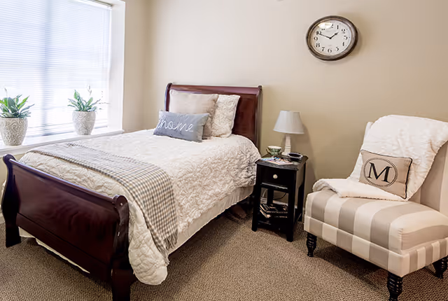 A cozy bedroom with a wooden sleigh bed dressed in white bedding and a gray throw blanket. There are two decorative pillows on the bed, one with the word 'home'. Next to the bed is a black nightstand with a lamp, a cup, and some books. A beige and white striped armchair with a white throw and a pillow with the letter 'M' sits nearby. A round wall clock hangs on the beige wall above the chair. Two potted plants are placed on the windowsill with blinds partially closed.
