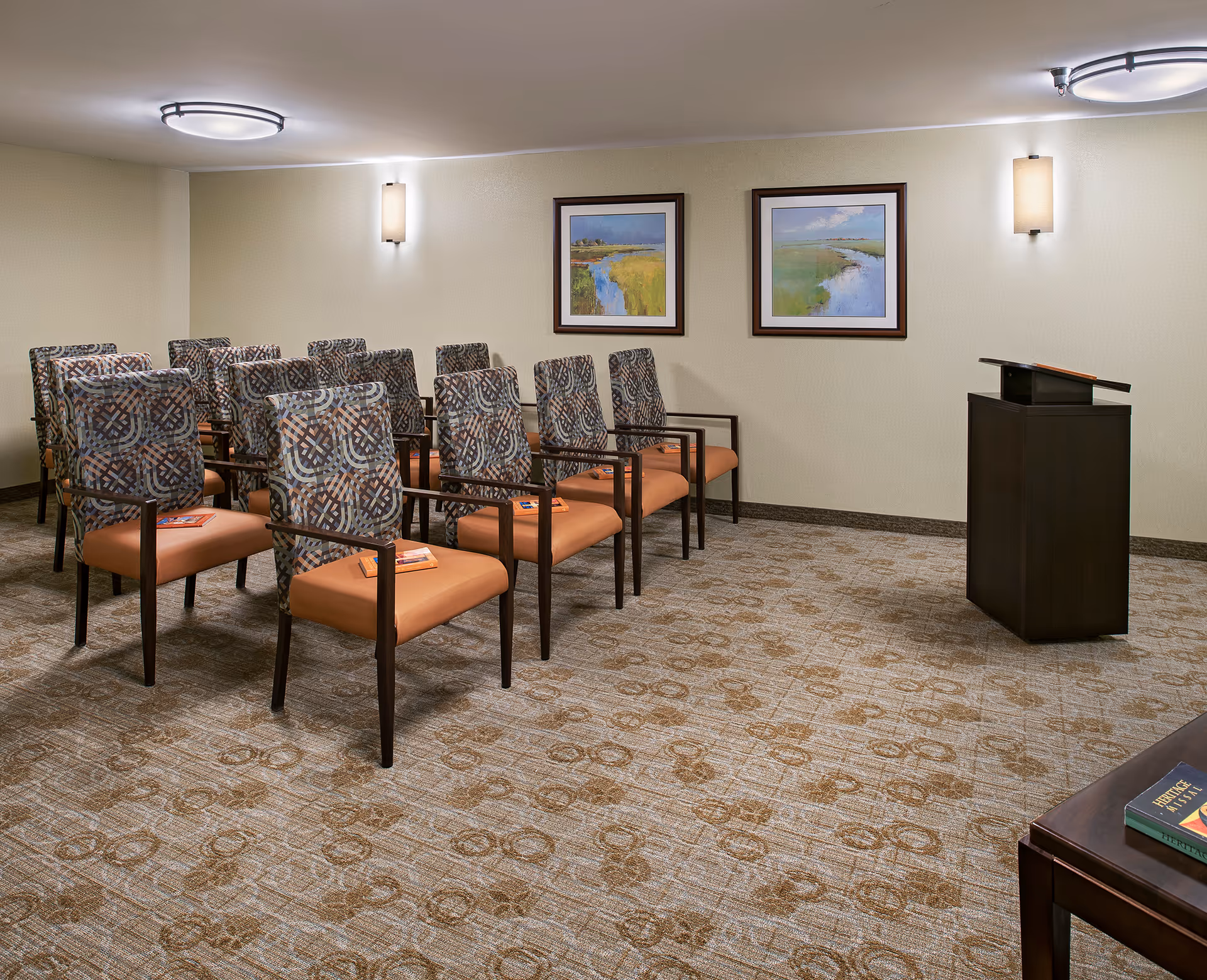 A small conference or meeting room with three rows of patterned chairs with orange seats facing a dark wooden podium. The room has beige walls with two framed landscape paintings and modern wall sconces. The carpet has a circular pattern in shades of brown.