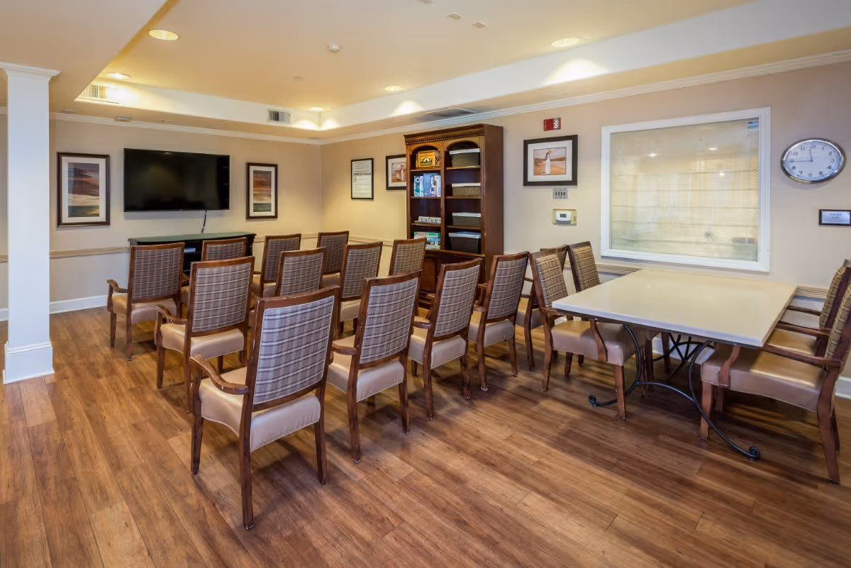 A meeting or activity room with rows of wooden chairs facing a wall-mounted flat screen TV. There is a wooden bookshelf with games and books, framed pictures on the walls, a clock, and a table with additional chairs on the right side. The room has wood flooring and soft lighting.
