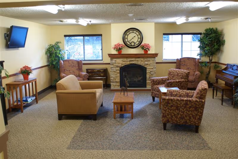 A cozy living room area with a stone fireplace centered on the back wall, flanked by two windows with blinds. There are two armchairs with floral patterns, one plain armchair, and a small wooden coffee table in the middle. A large clock hangs above the fireplace, and there are potted plants and flowers on either side. A flat-screen TV is mounted on the left wall, and a piano is visible on the right side of the room.