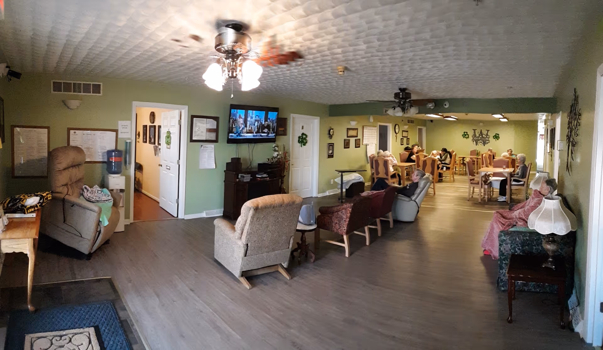 Communal living and dining area with armchairs and dining tables, a wall-mounted TV, and several seated residents.