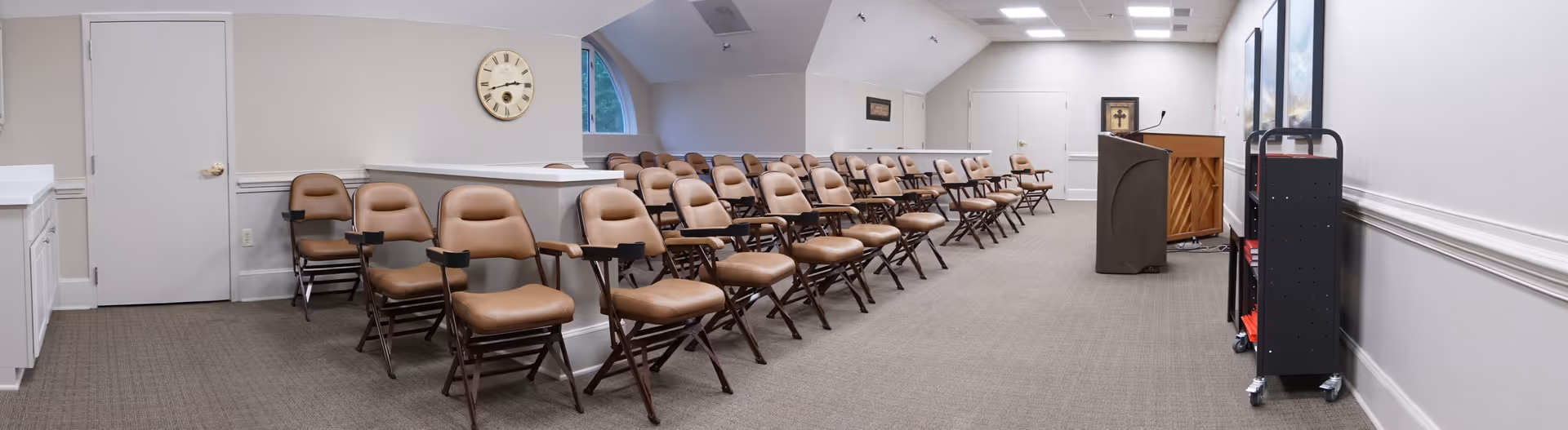 A large room set up with multiple rows of brown cushioned chairs with armrests, arranged facing a podium and a piano at the front. The room has beige walls, a carpeted floor, a clock on the wall, and fluorescent ceiling lights. There is a black shelving unit on the right side near the podium.