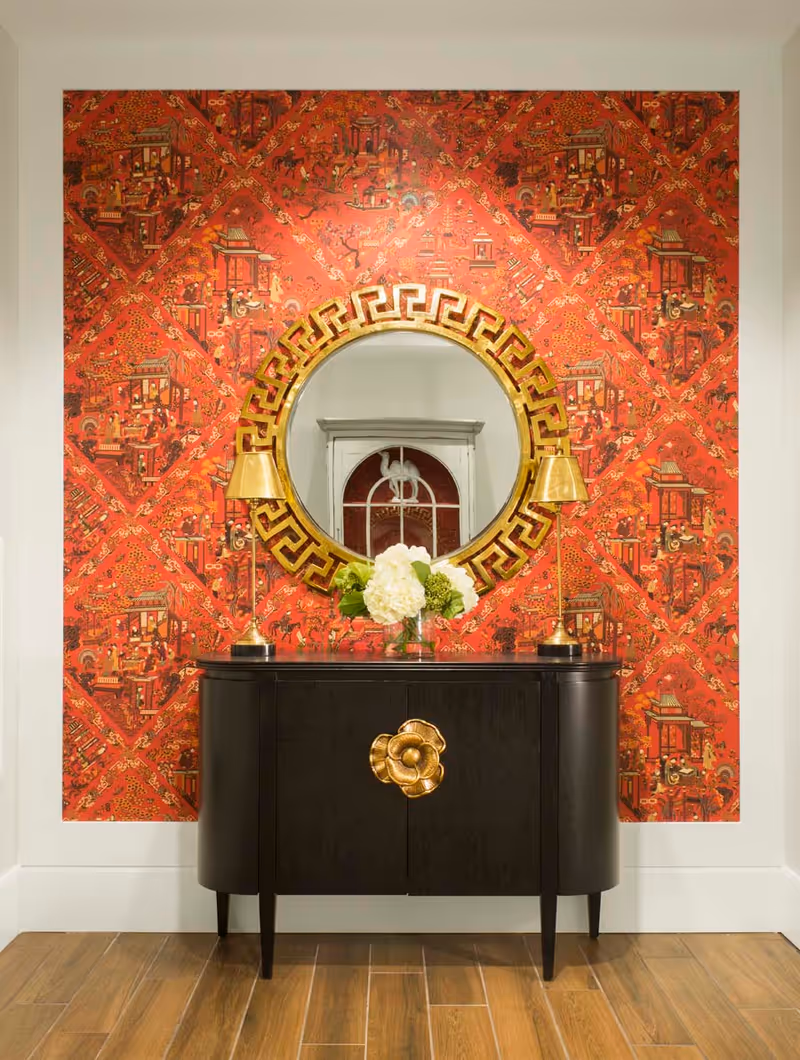 A decorative interior wall with red patterned wallpaper featuring Asian-inspired designs. A round mirror with a gold geometric frame is centered on the wall above a black console table with a large gold flower-shaped handle. Two gold lamps flank a vase of white and green flowers on the table. The floor is wooden.