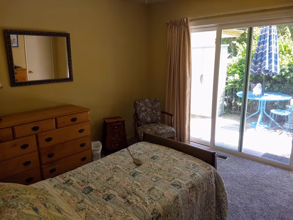 A bedroom with a bed covered in a patchwork quilt, a wooden dresser with a mirror above it, a small wooden nightstand, and a floral upholstered chair. There is a sliding glass door with beige curtains leading to an outdoor patio with a blue table, chairs, and a striped umbrella.