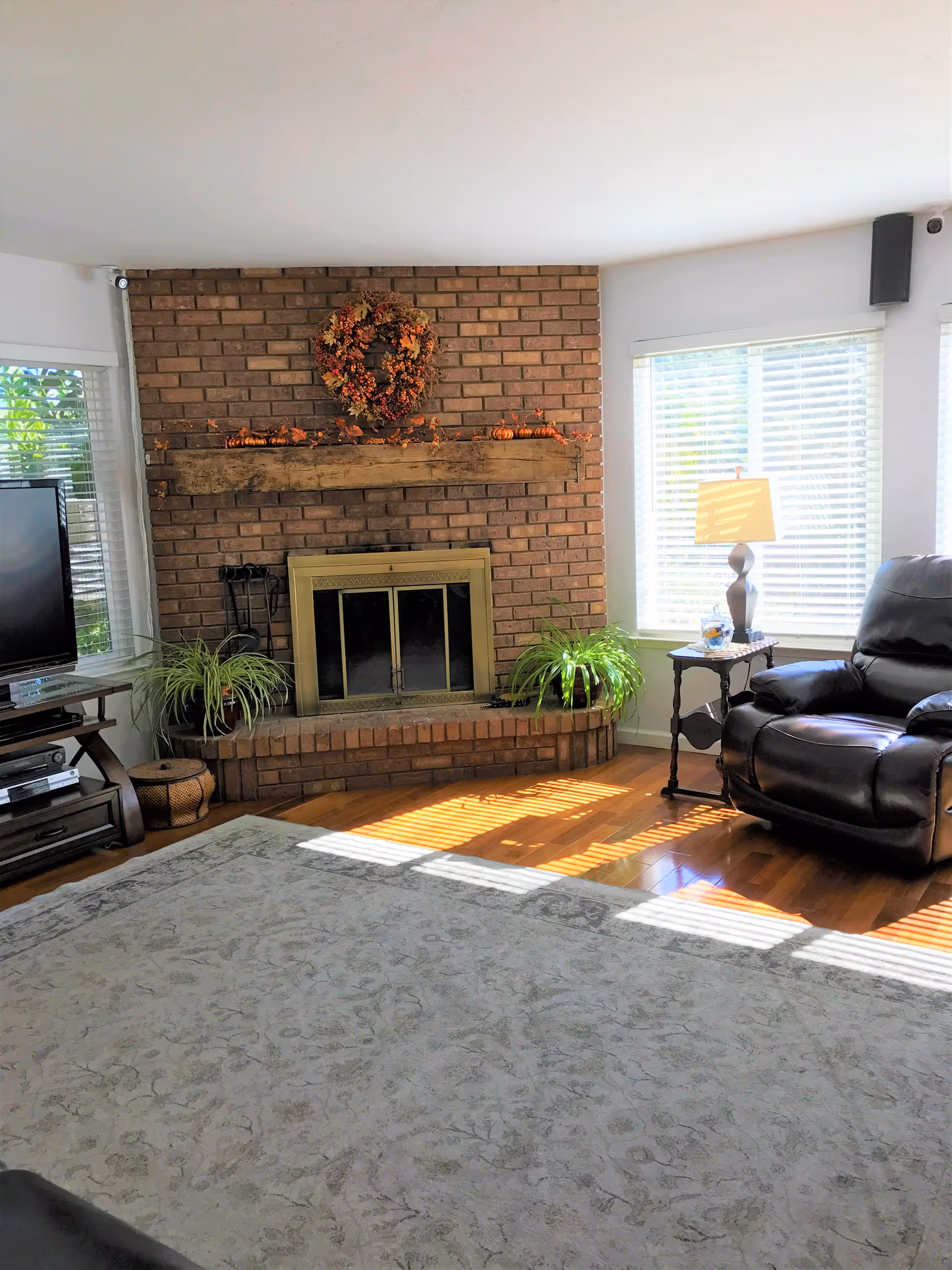 Sunlit living room with a brick fireplace and wooden mantel decorated with an autumn wreath, a leather recliner, TV, plants, and a large area rug.