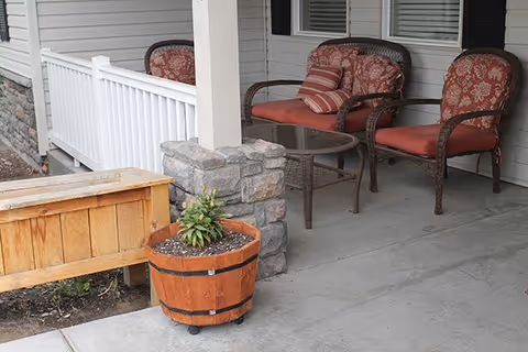 A covered outdoor patio area with three cushioned wicker chairs and a glass-top coffee table. There is a wooden planter with a small plant and a wooden bench nearby. The patio is adjacent to a building with white siding and windows with blinds.