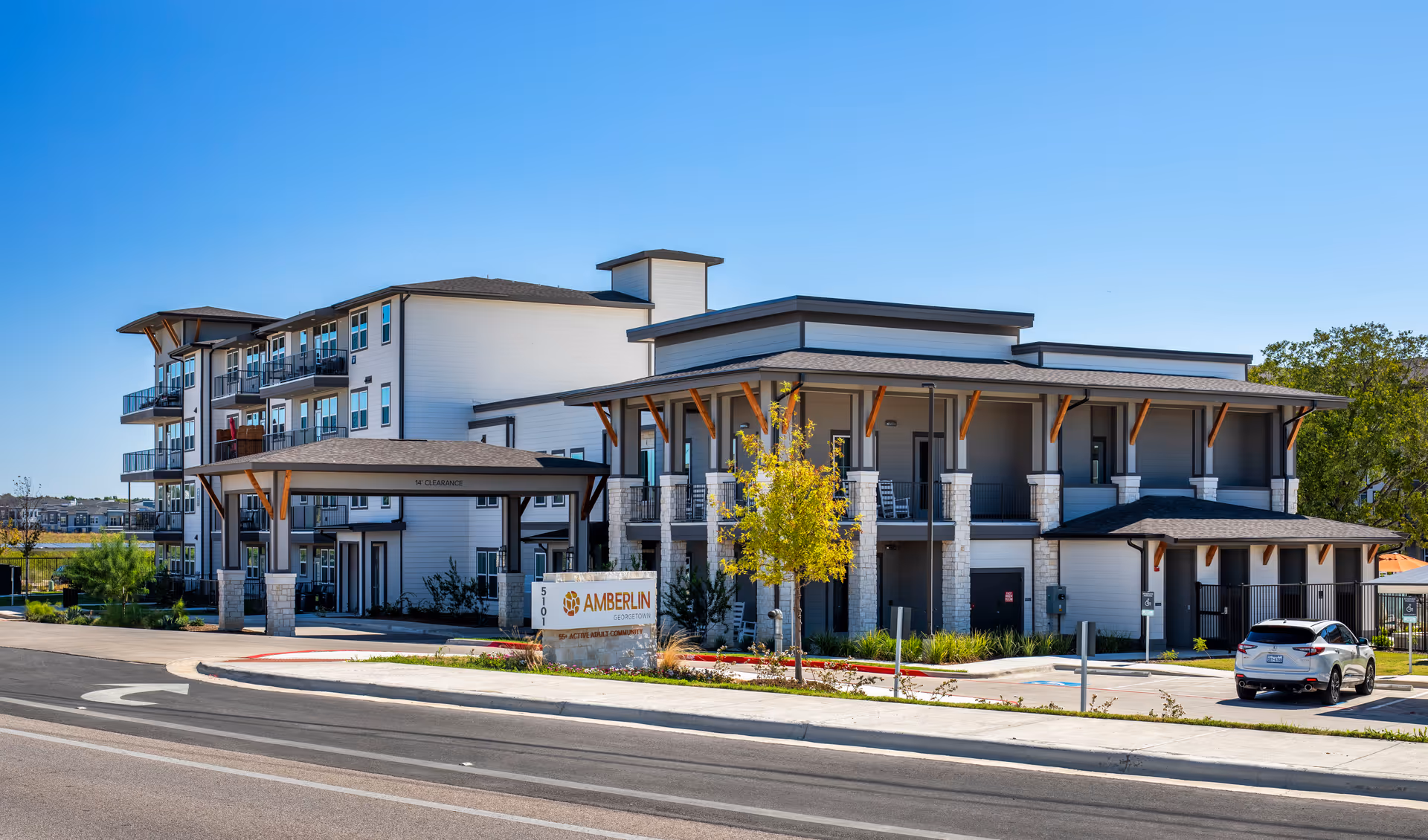 Exterior view of Amberlin Georgetown, a modern multi-story senior living facility with balconies, a covered entrance, and a parking area with a car. The building is surrounded by landscaping and a clear blue sky.