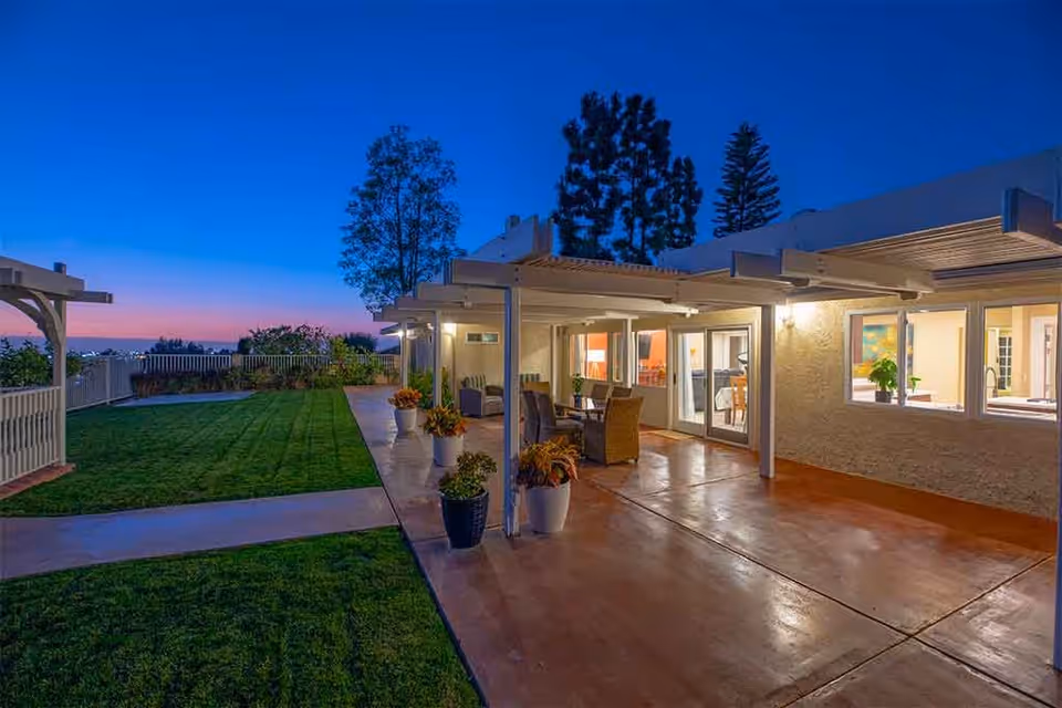 Covered patio with outdoor seating beside a manicured lawn and illuminated building at dusk.