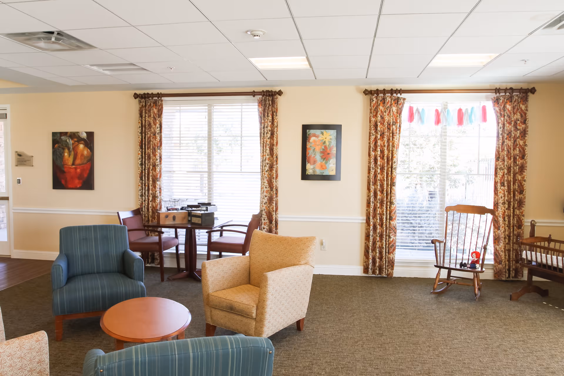 A bright and cozy common area in an assisted living facility featuring comfortable armchairs, a small round table, a wooden rocking chair, and a crib. The room has large windows with floral curtains, framed artwork on the walls, and a small table with chairs near the window.