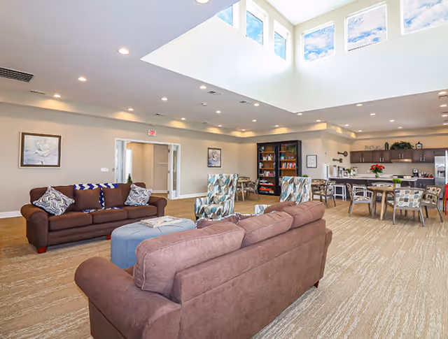 A spacious and well-lit common area in a senior living facility featuring two brown sofas with patterned cushions, two patterned armchairs, a blue ottoman, several dining tables with chairs, a bookshelf, and a kitchen area in the background. Large windows near the ceiling allow natural light to fill the room.