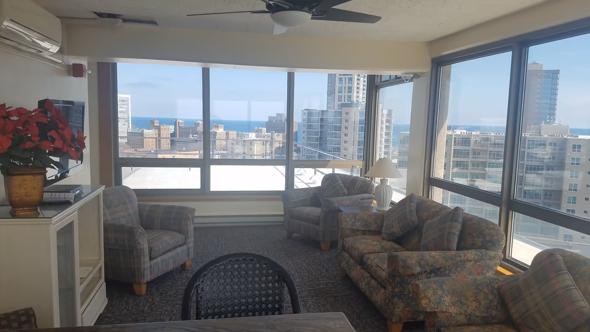 A cozy living room with large windows offering a panoramic view of city buildings and a distant body of water. The room features a floral-patterned sofa, two plaid armchairs, a side table with a lamp, a TV mounted on the wall, and a cabinet with a potted plant on top.