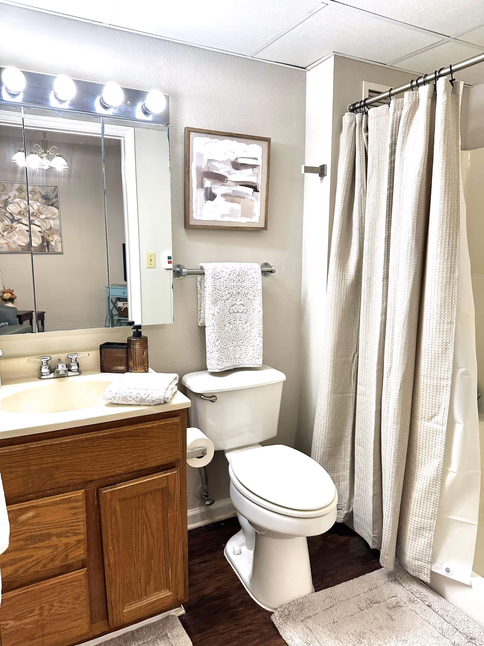 A clean and well-lit bathroom featuring a white toilet, a wooden vanity with a sink, a large mirror with lights above it, a towel rack with a white patterned towel, a shower with a beige curtain, and a framed abstract painting on the wall.