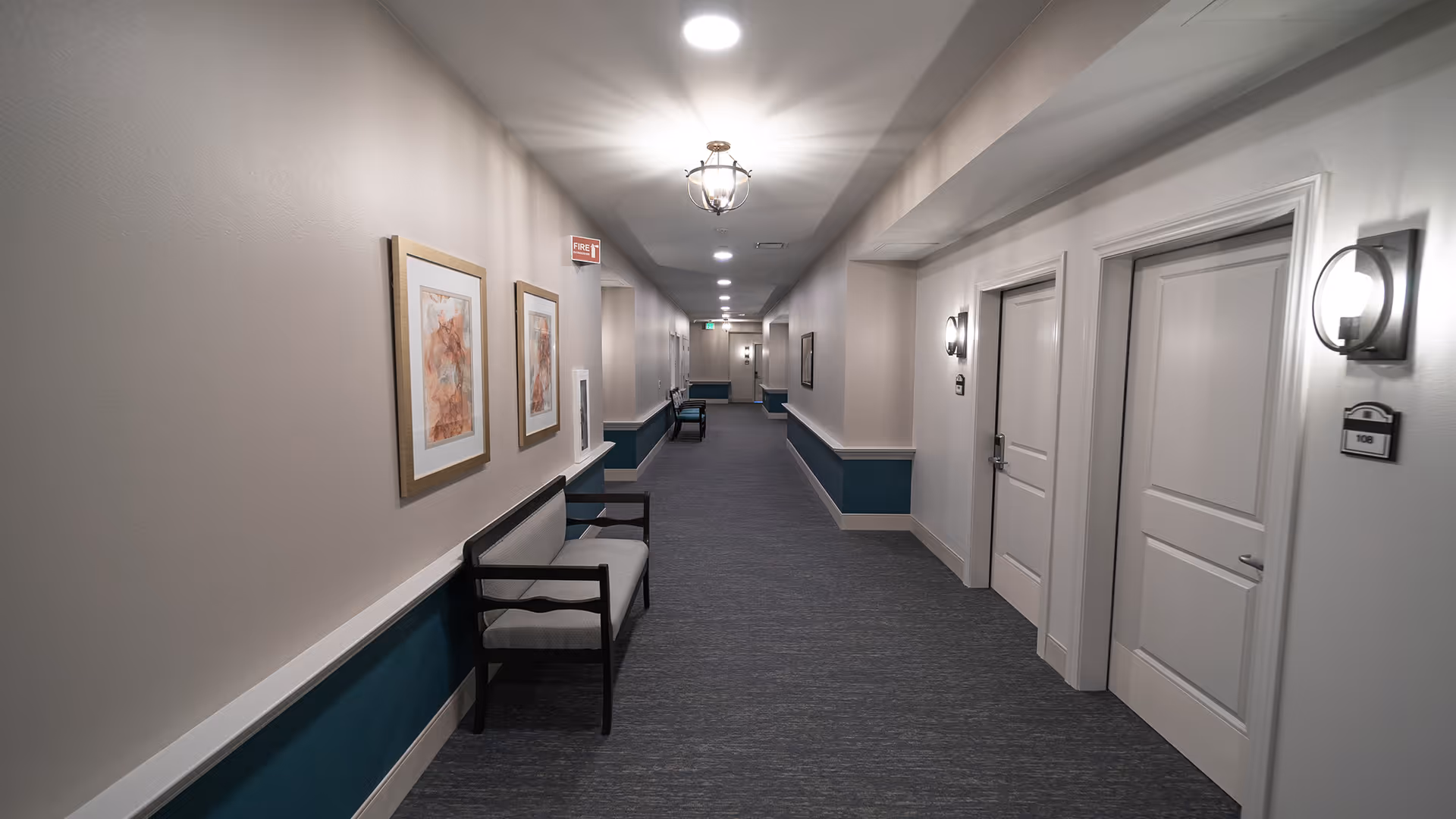 A well-lit, clean hallway in a senior living facility with light-colored walls, dark blue wainscoting, and carpeted flooring. There are framed artworks on the left wall and several benches along the corridor. Multiple white doors with room numbers are visible on the right side, and ceiling lights illuminate the space.