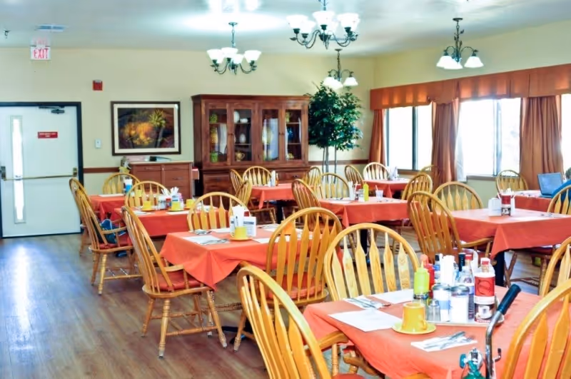 A dining room with multiple wooden tables covered with red tablecloths and wooden chairs. Each table is set with condiments, utensils, and cups. The room has large windows with orange curtains, a wooden cabinet with glass doors, a potted plant, and ceiling chandeliers providing light.