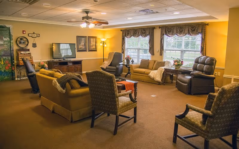 A cozy living room in a senior living facility with multiple armchairs and sofas arranged around a central area. The room features a ceiling fan with lights, a television on a wooden stand, large windows with valances letting in natural light, and decorative elements like framed pictures, flowers, and a floor lamp.