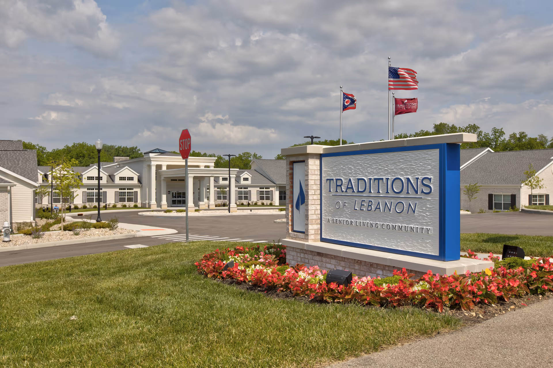 Exterior view of Traditions of Lebanon senior living community with a large sign in the foreground surrounded by flowers, a driveway, and the building entrance in the background under a partly cloudy sky.