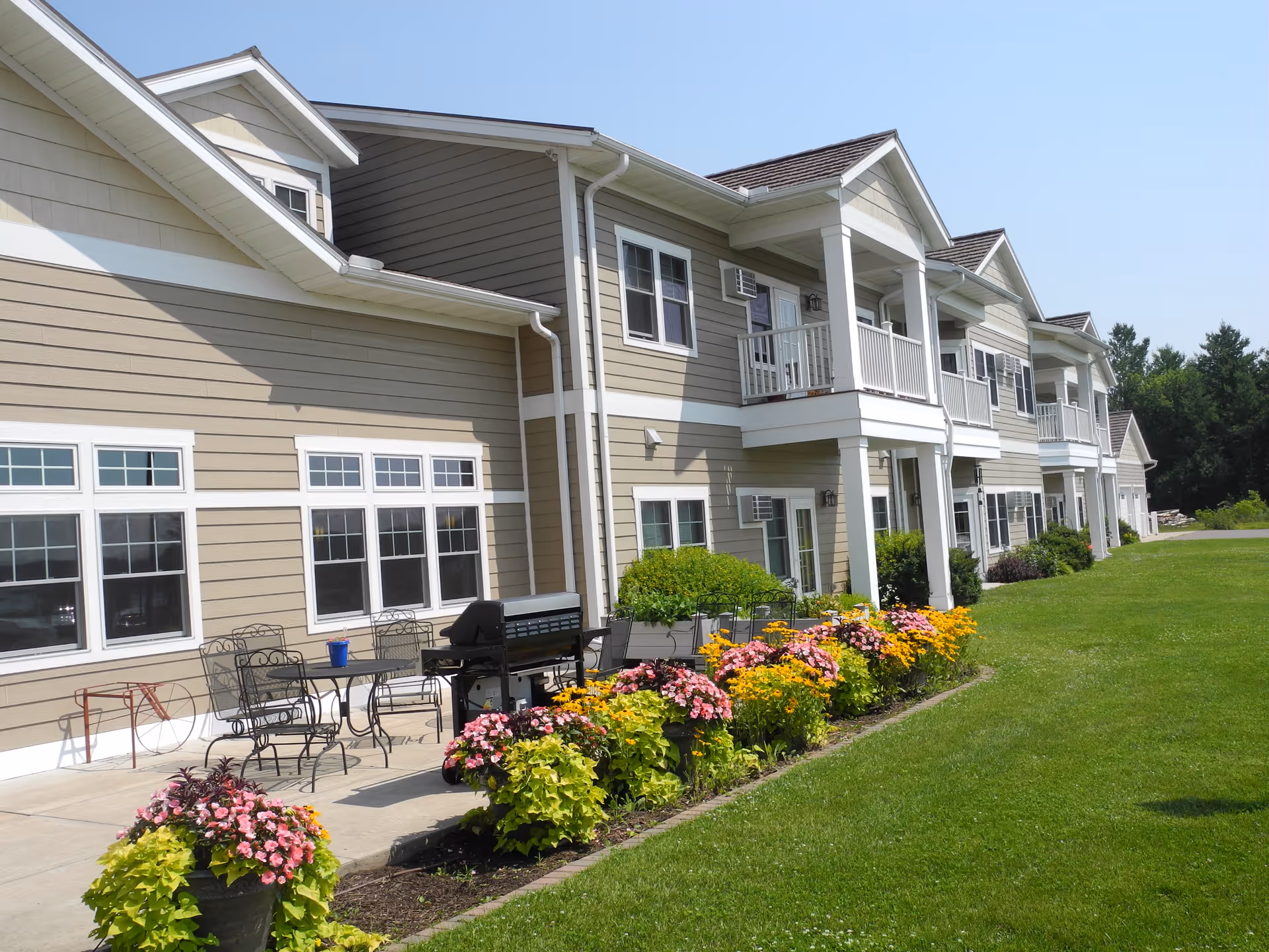 Exterior view of a two-story senior living facility building with beige siding and white trim. The building features balconies on the upper floor and a patio area with metal chairs, tables, and a grill. There are colorful flower beds along the edge of the patio and a well-maintained green lawn extending to the right. Trees are visible in the background under a clear blue sky.